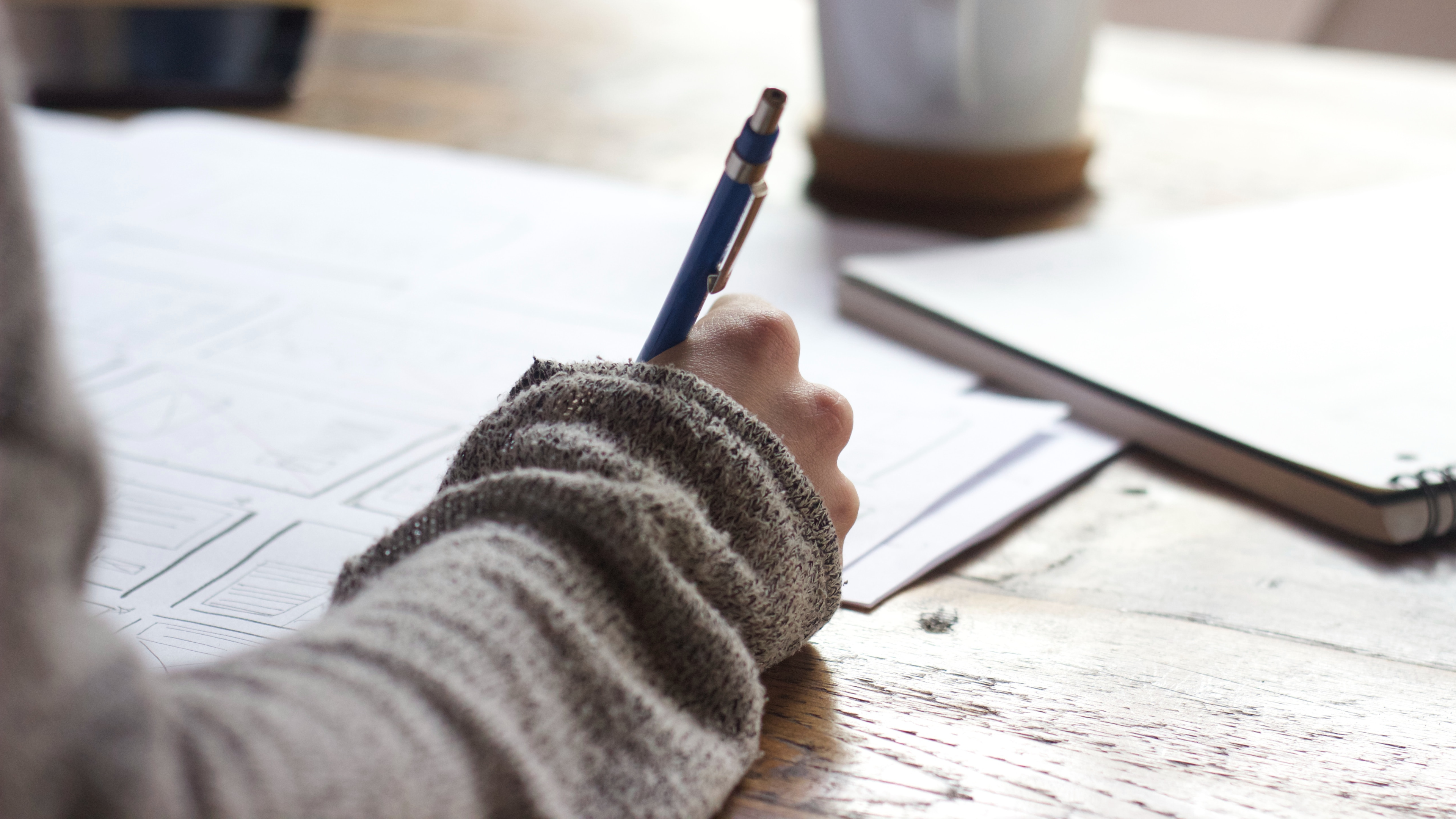 An image of a women's arm in a comfortable sweater writing notes on a desk. Photo by https://unsplash.com/@uns__nstudio?utm_content=creditCopyText&utm_medium=referral&utm_source=unsplash Unseen Studio on https://unsplash.com/photos/person-writing-on-brown-wooden-table-near-white-ceramic-mug-s9CC2SKySJM?utm_content=creditCopyText&utm_medium=referral&utm_source=unsplash